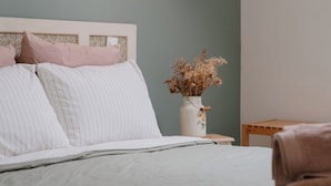 Cama bem arrumada com roupa de cama listrada, abajur, um vaso com flores secas e um quadro na parede.