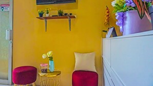A room with a yellow wall, a TV mounted above a shelf with plants, a small round table with a vase, two red chairs, and a white cabinet. - OYO 92292 Nadjla Property Cibubur Village (Depok)