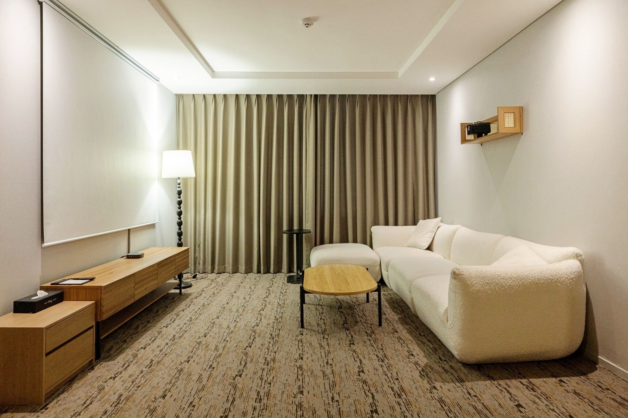 A hotel room with a wooden desk, a small round table, a white sofa, a lamp, a television, and a large window with curtains.
