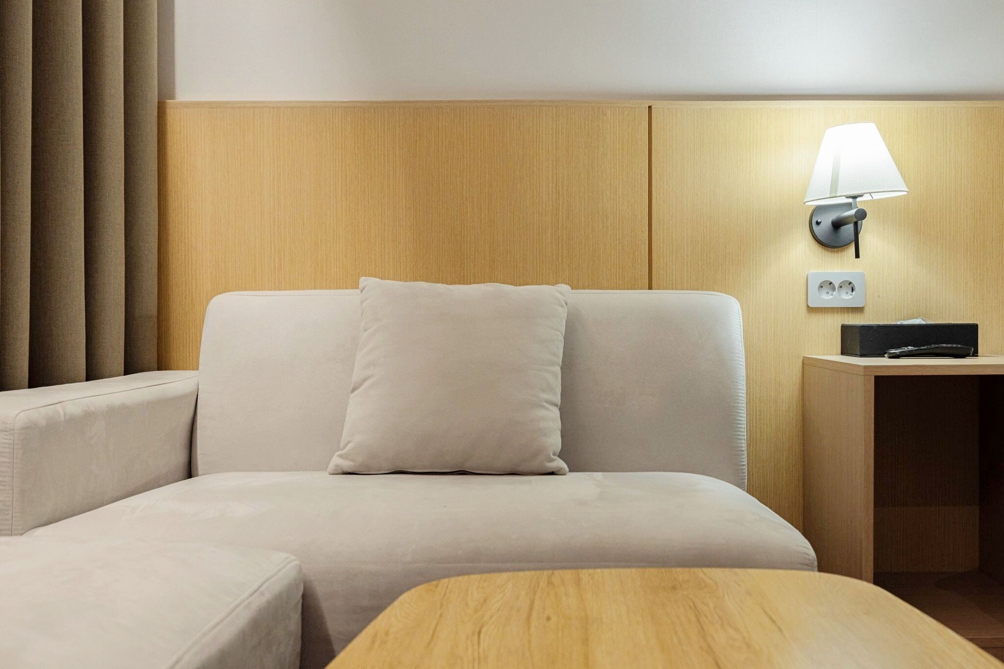 A hotel room with a sofa, a wooden side table, a lamp, and a power outlet.