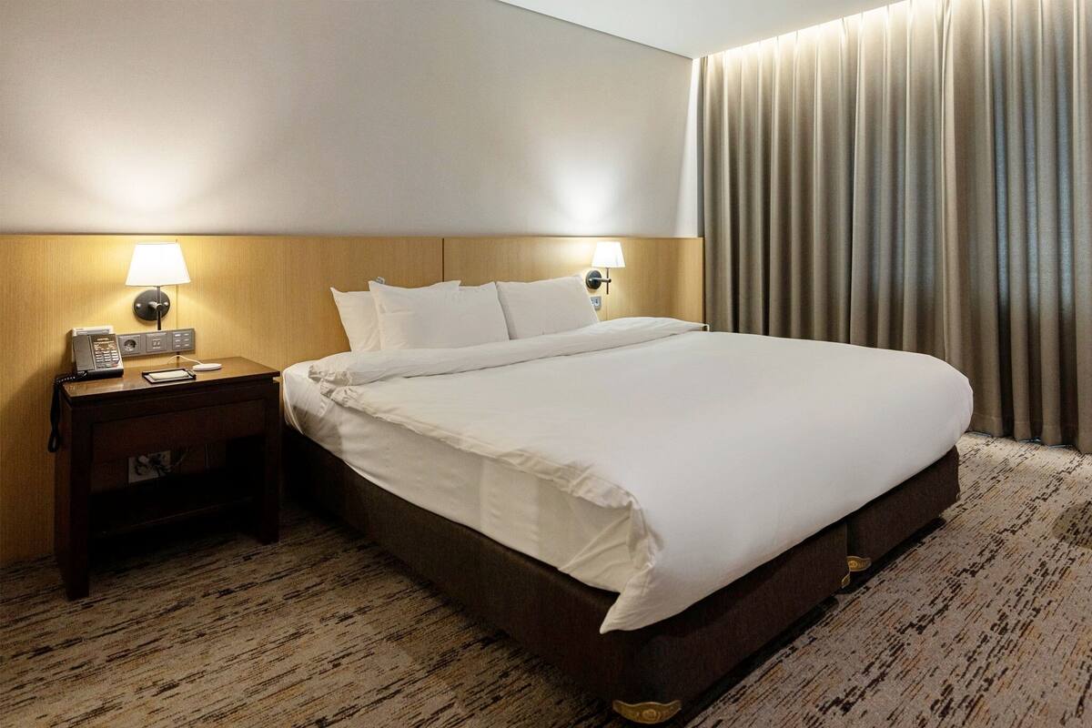 Una habitación de hotel con una cama grande, dos lámparas de noche, una mesita de noche con un teléfono y un bloc de notas, y una cortina en la ventana.