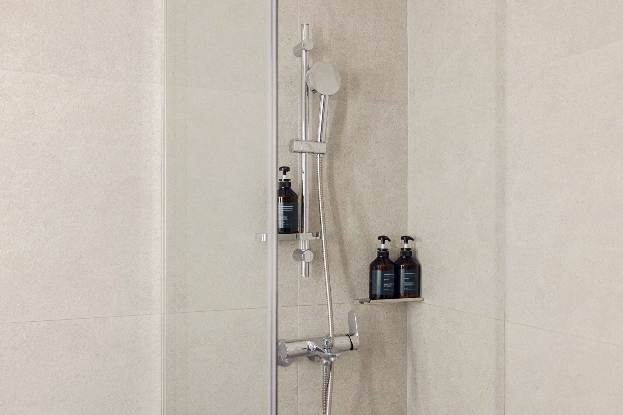 A shower with a handheld showerhead, a wall-mounted faucet, and a shelf with two bottles.