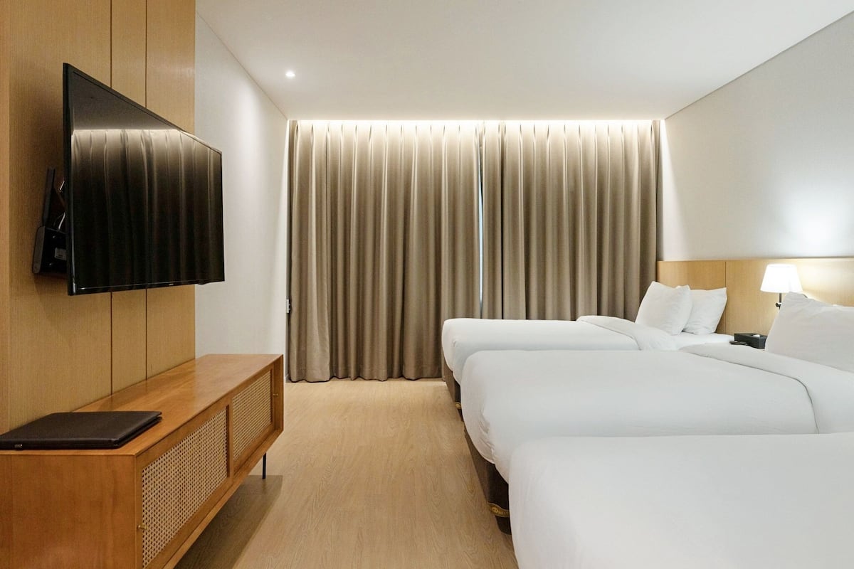 Habitación de hotel con televisión de pantalla plana en la pared, una mesita de madera con una computadora portátil, dos camas con sábanas blancas, un ventanal con cortinas y una lámpara de noche.