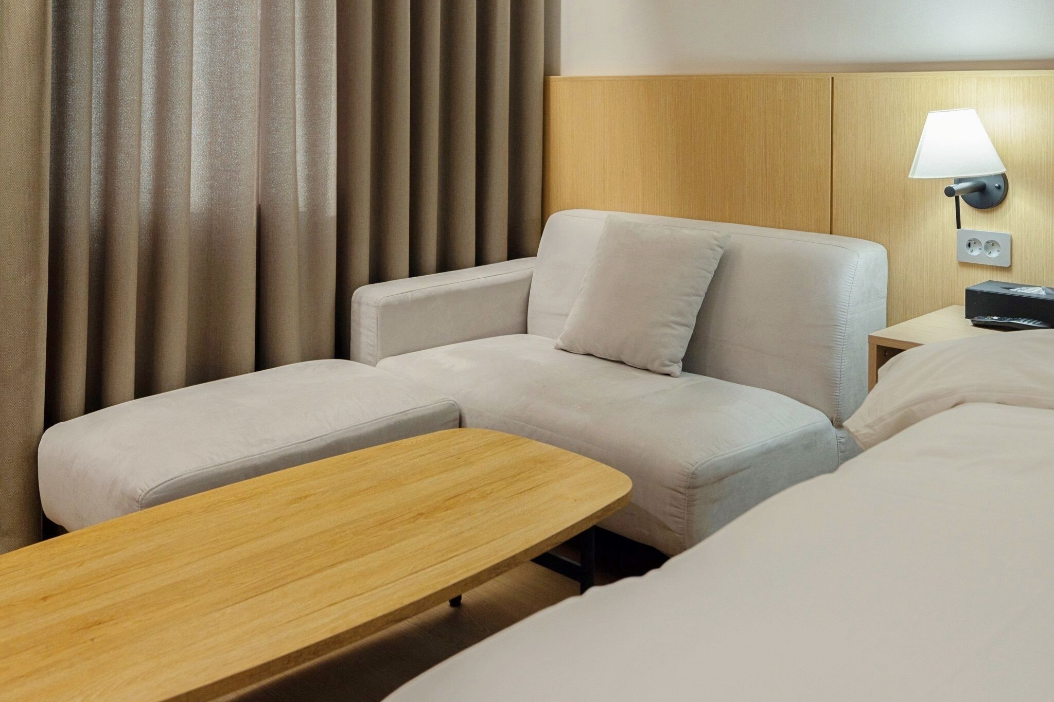 Quarto de hotel com sofá, mesa lateral de madeira, luminária e tomada na parede.
