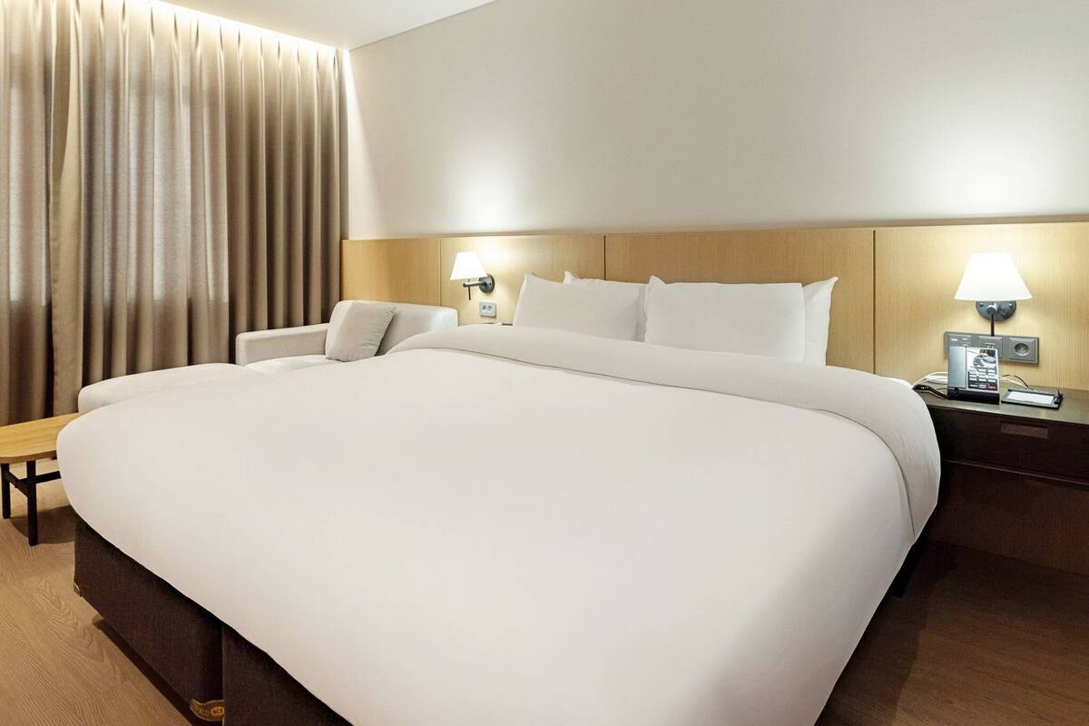 Habitación de hotel con una cama grande, mesitas de noche con lámparas, cabecera de madera y una ventana con cortinas.