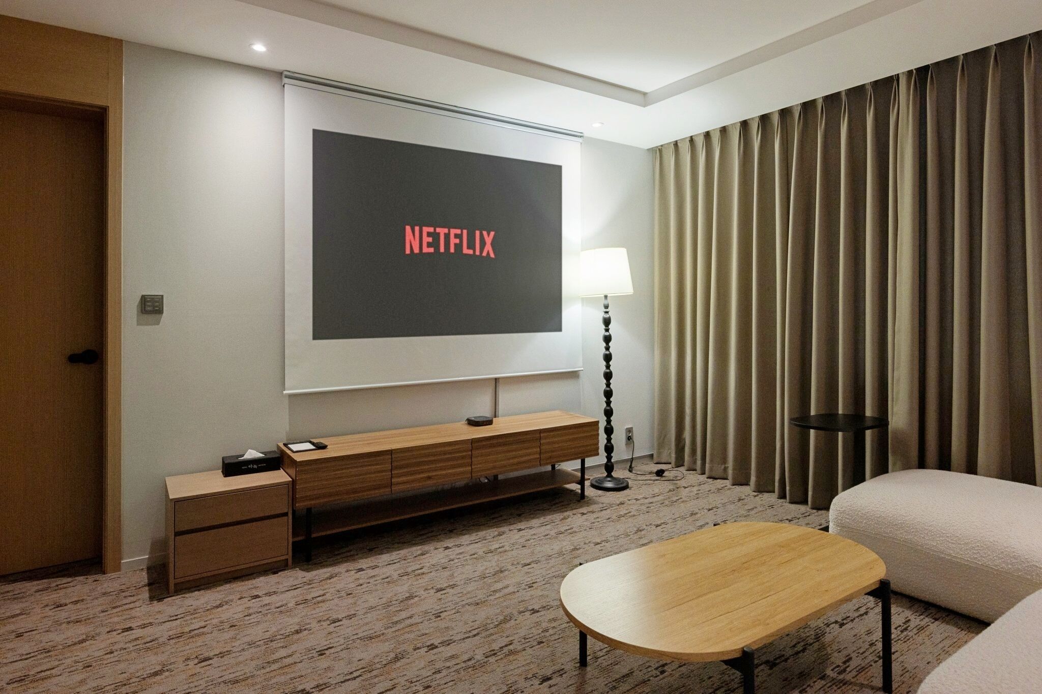 Een hotelkamer met een grote flatscreen-tv waarop 'NETFLIX' wordt weergegeven, een houten bijzettafeltje, een lamp, een bank en een vloerbedekking.