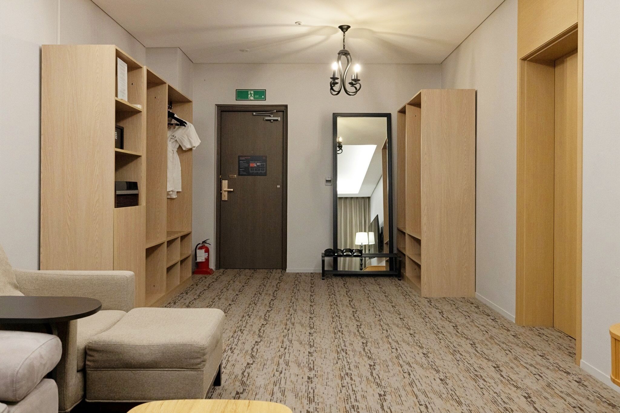 A hotel room with a sofa, a small table, a wardrobe, a mirror, and a door with an emergency exit sign.