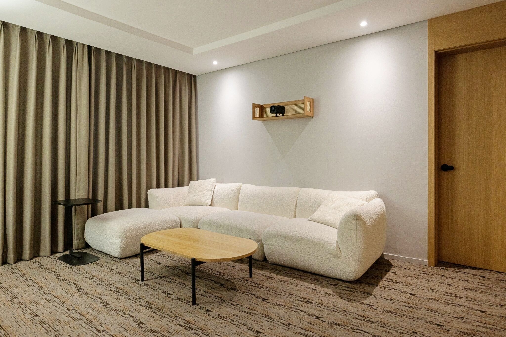 Un salon moderne avec un canapé d’angle, une petite table ronde, un téléviseur fixé au mur et une porte.