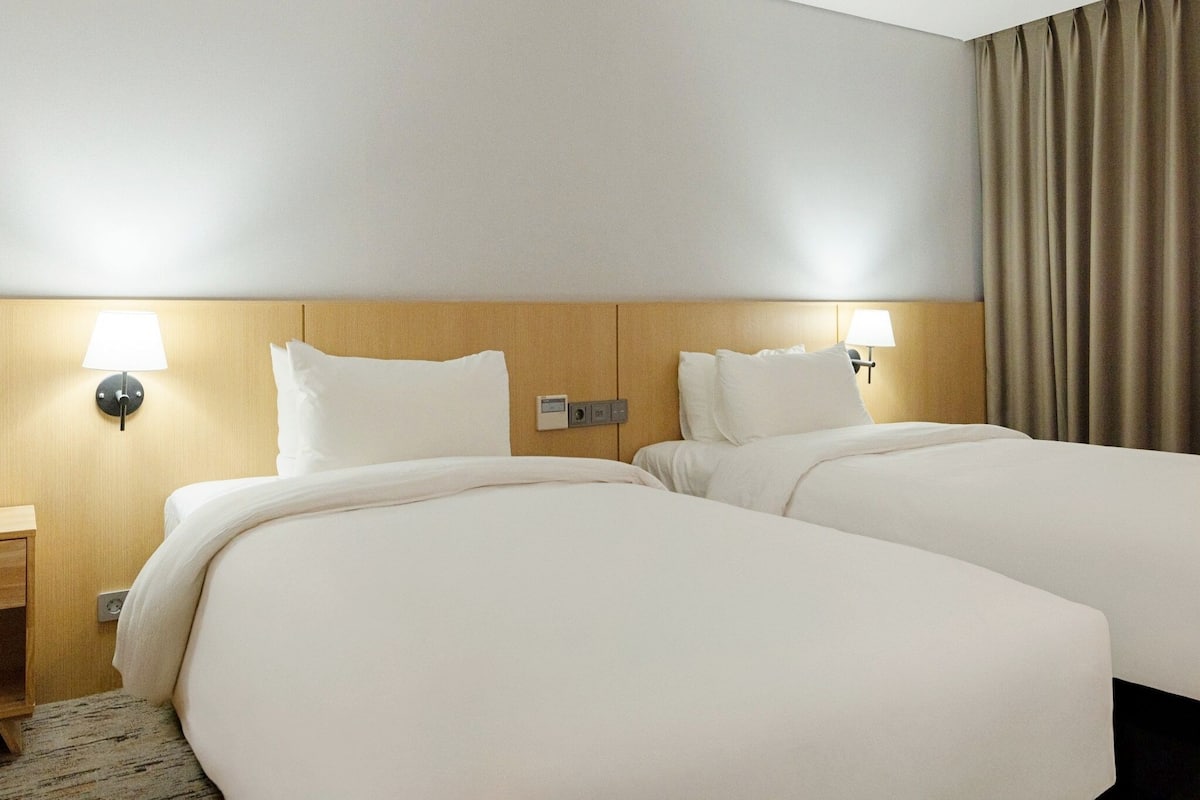 Habitación de hotel con dos camas, lámparas de noche, cabecera de madera, mesitas de noche y una cortina.