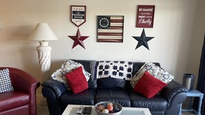 A living room with a black leather sofa, red and white patterned pillows, a white coffee table with remote controls and a basket of yarn balls, and wall decorations including a star, a wreath, and two patriotic-themed signs.