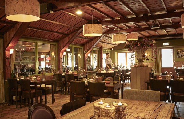 A restaurant with wooden beams, hanging lamps, and a bar area. Tables are set with candles and wine glasses.