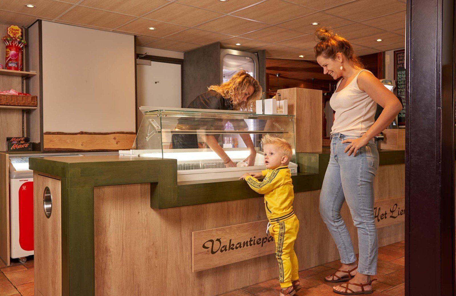 A wooden counter with a glass display case, a sign reading "Vakantiepau," and a red appliance in the background.