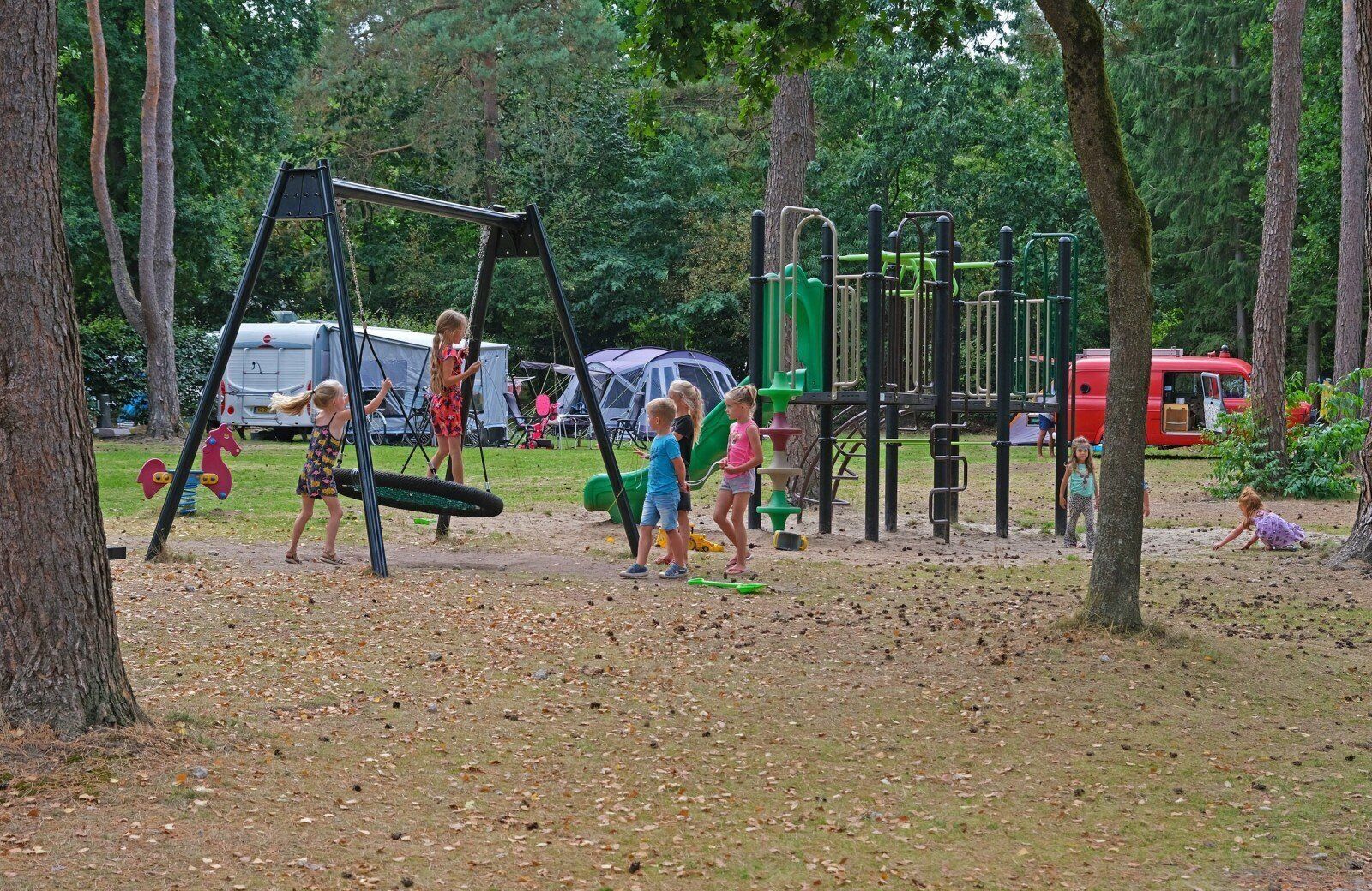 A playground with swings and climbing structures, surrounded by trees and camping tents in the background.