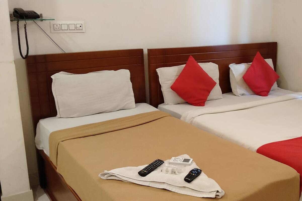 Quarto de hotel com duas camas, cada uma com roupa de cama branca e almofadas vermelhas. Há um cobertor castanho no chão, e um par de controles remotos sobre ele.