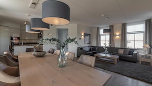 Ein modernes Wohnzimmer mit einem Holzesstisch, Ledersesseln und einer großen Vase mit Grünpflanzen. Im Hintergrund befindet sich eine Couch und ein Couchtisch.