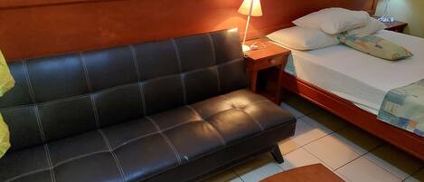 A hotel room with a black leather sofa, a wooden nightstand with a lamp, a bed with white bedding and a patterned pillow, and a tiled floor.