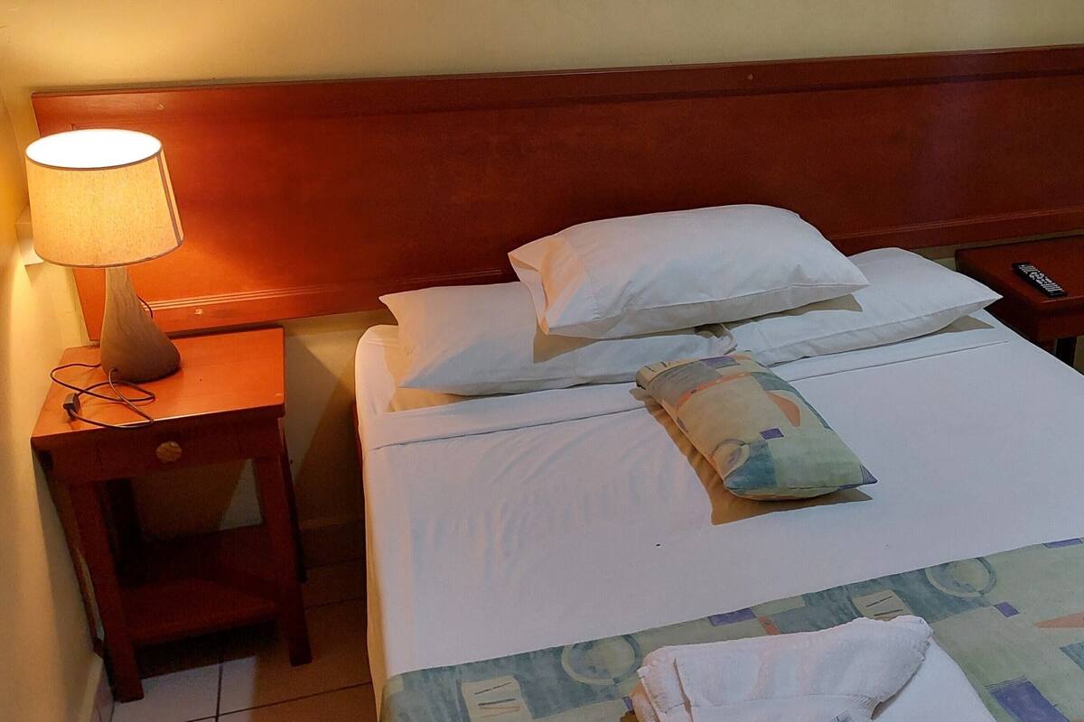 A hotel room with a wooden headboard, two beds with white bedding, a bedside table with a lamp, and a small nightstand with a remote control.