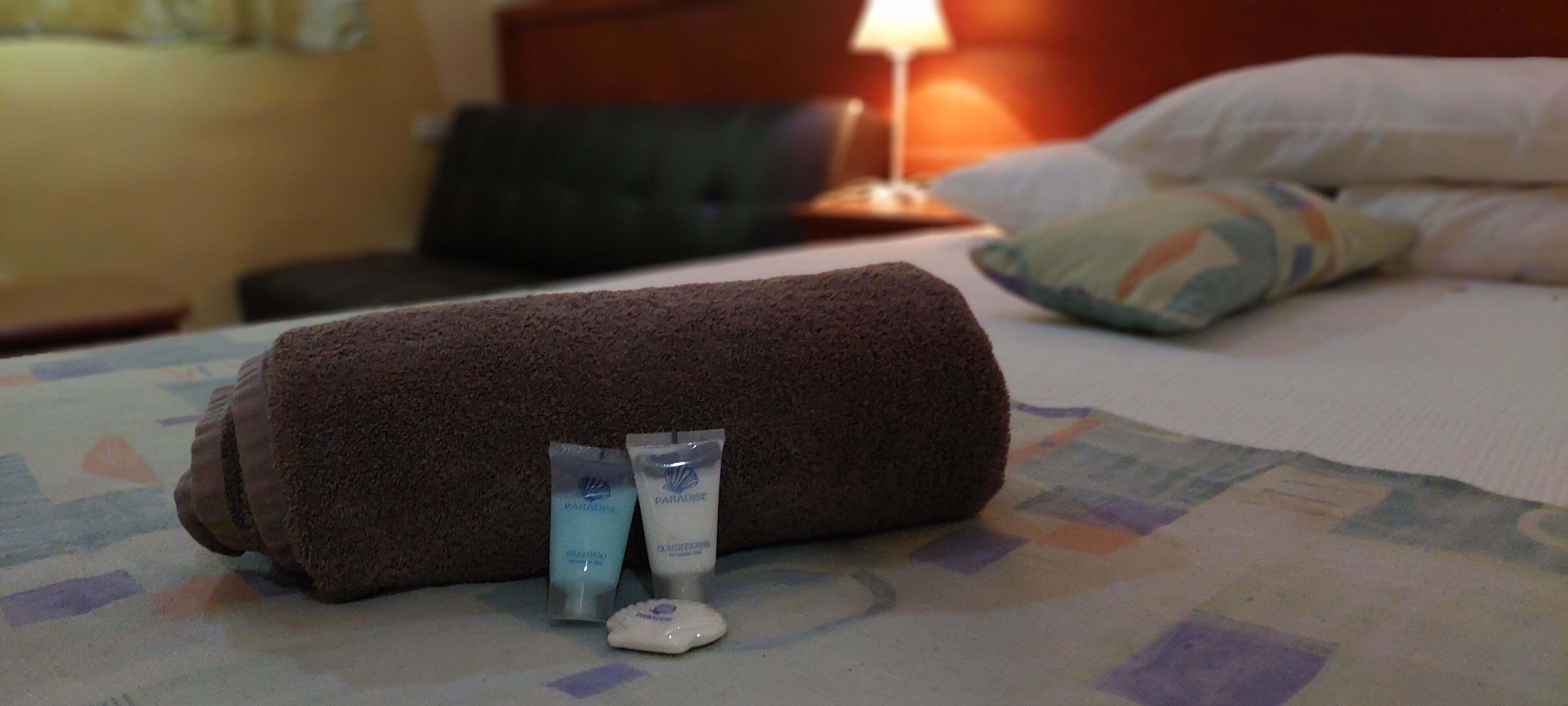 A neatly made bed with a rolled towel, two bottles of shampoo, and a bar of soap on a patterned bedspread.