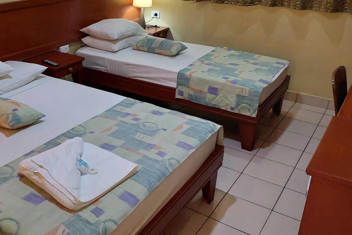 A hotel room with two beds, each with a pillow and a blanket. There is a nightstand with a lamp and a remote control.