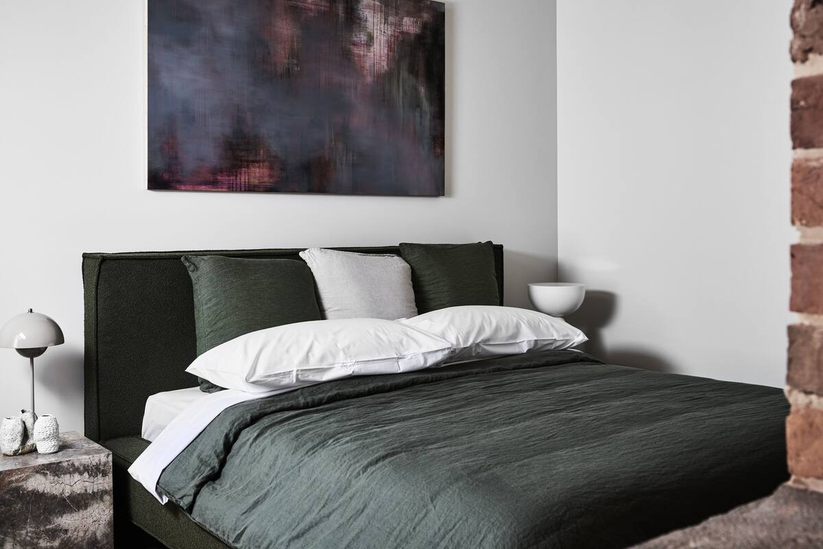 Un dormitorio moderno con una cama verde oscuro, almohadas blancas y un cuadro abstracto grande en la pared.