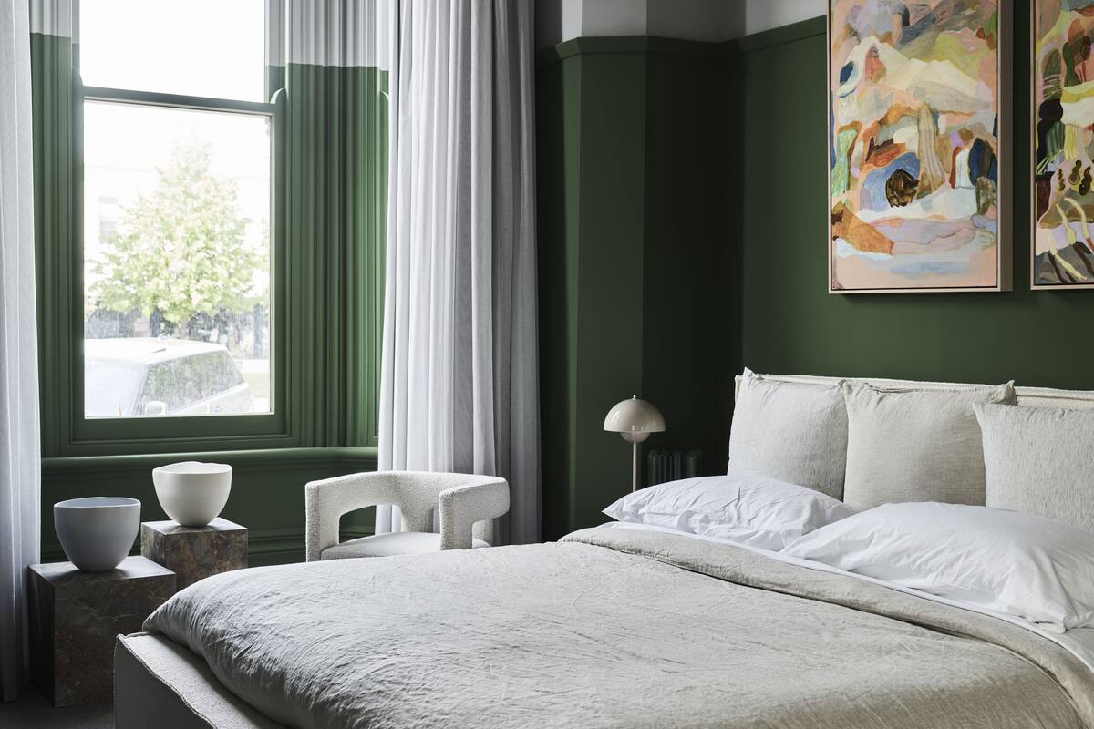 Un dormitorio con cama, una silla, una ventana con cortinas y un cuadro en la pared.