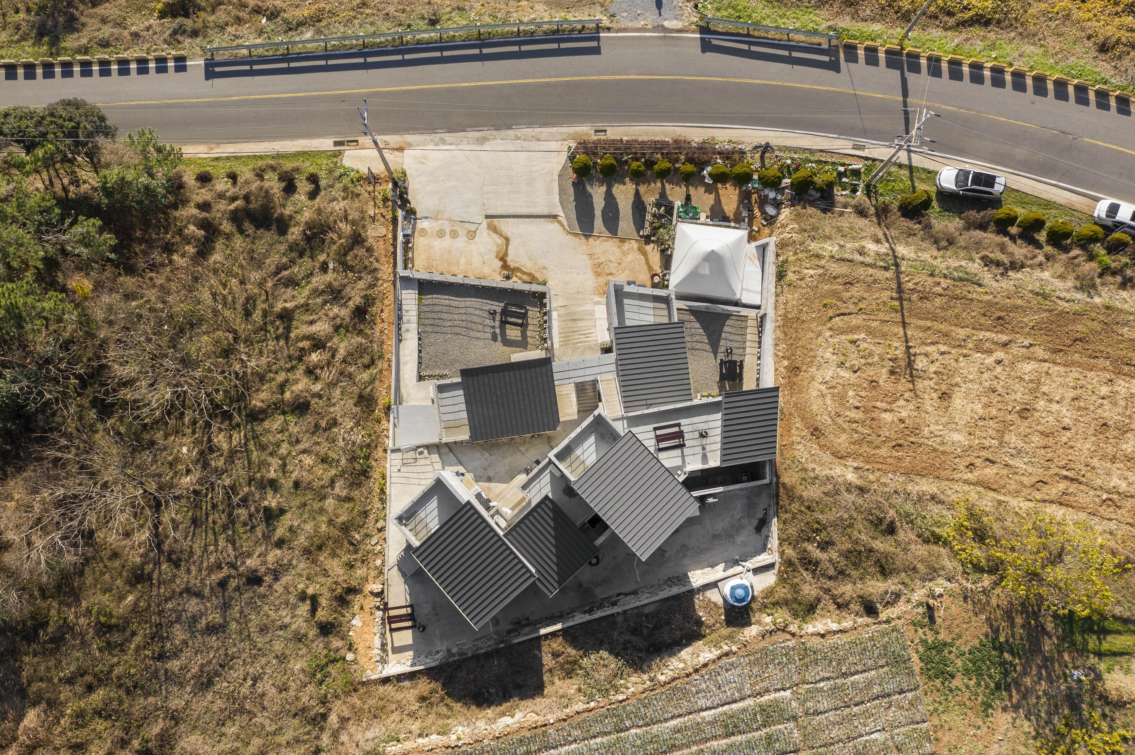 An aerial view of a building with a sloped roof, a paved driveway, and a road with a guardrail.