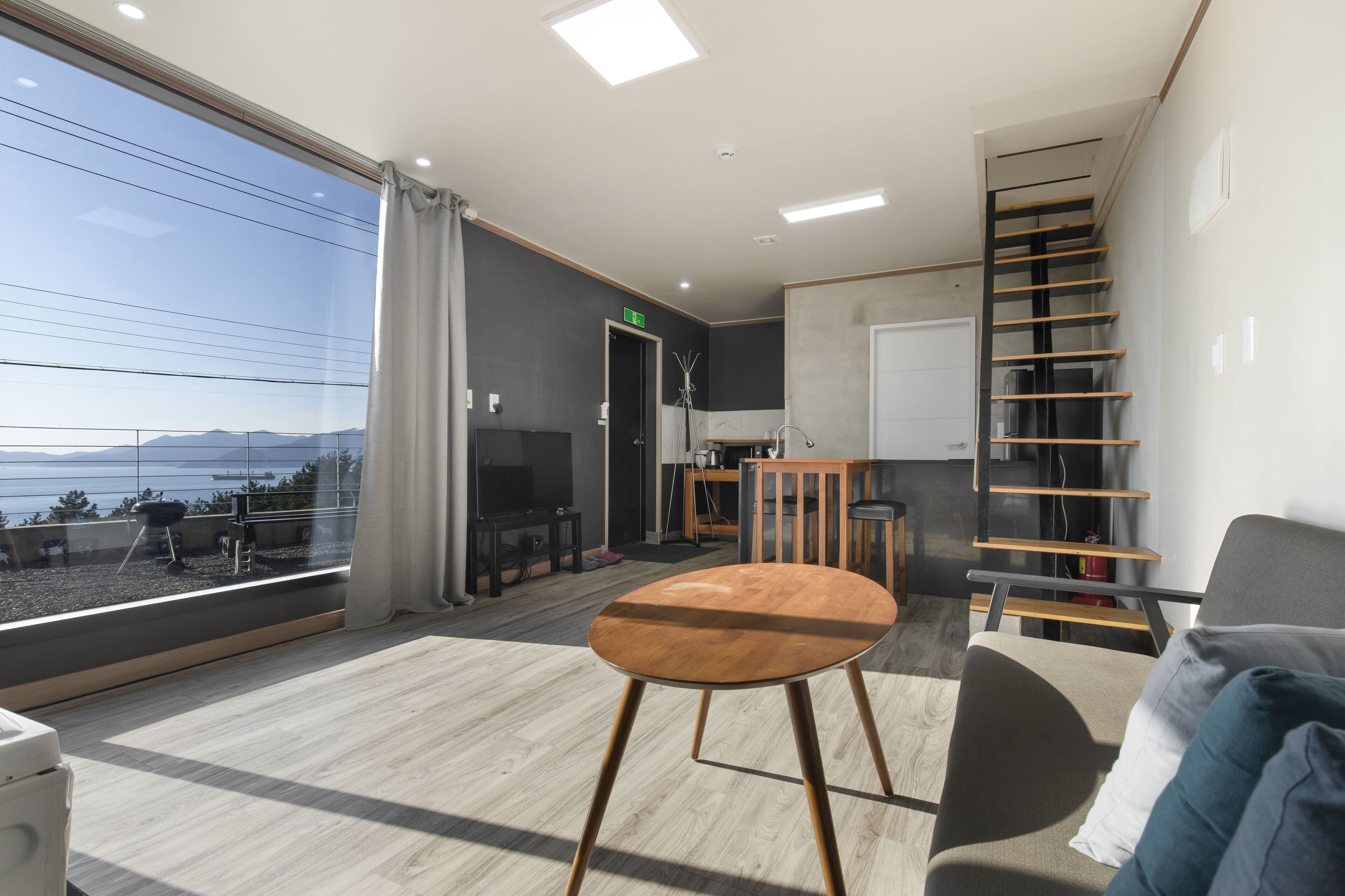 A modern living room with a large window, a wooden table, a sofa, and a staircase.