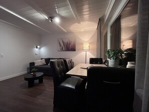 A modern living room with a dark leather sofa, a wooden coffee table, and a dining table with chairs. There is a floor lamp and a potted plant on the table.
