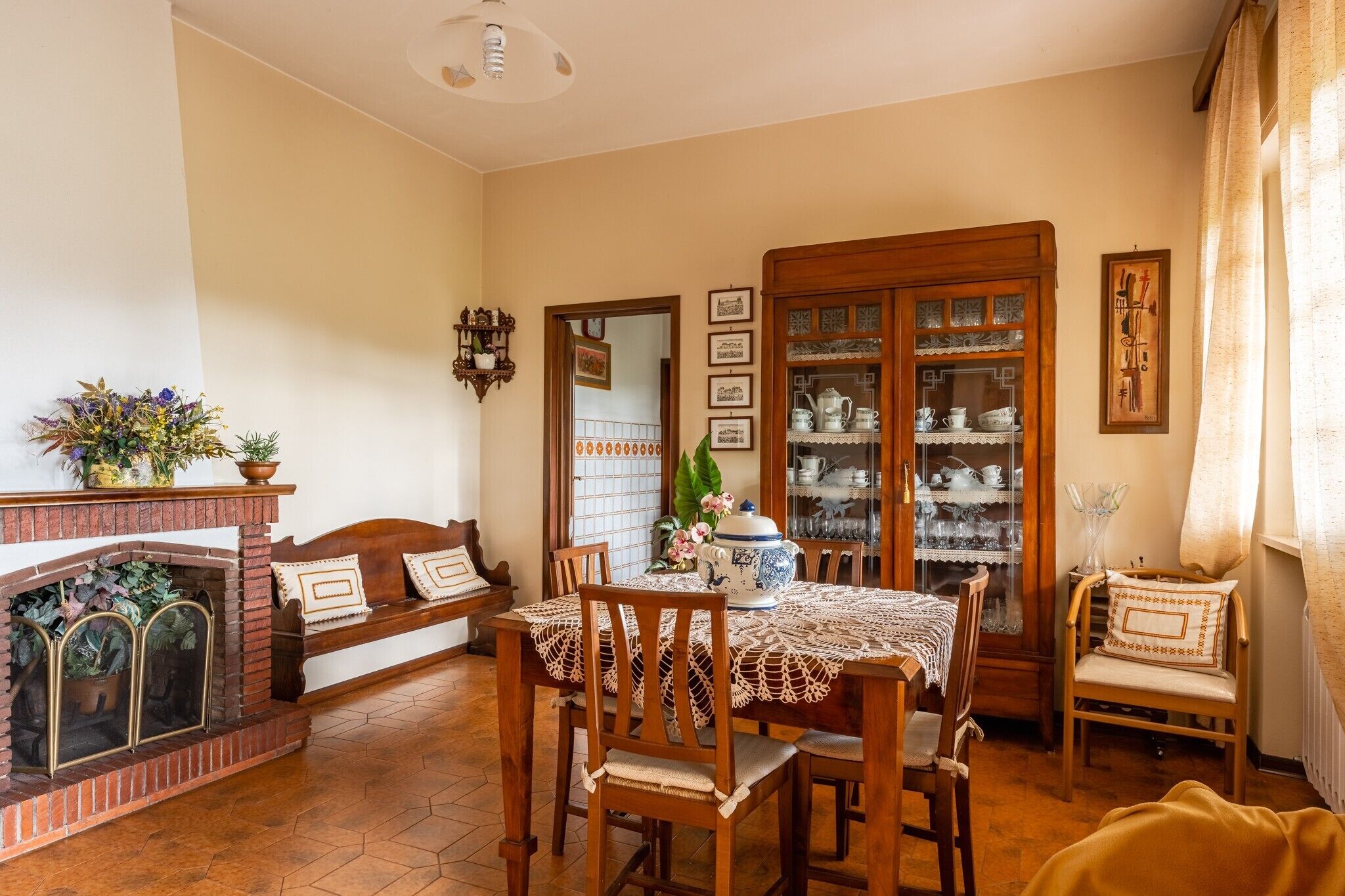 A dining area with a brick fireplace, wooden chairs, a table with a lace tablecloth, and a china cabinet with glass doors.