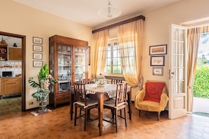 Un comedor con una mesa de madera, sillas, un armario con puertas de vidrio, un jarrón con flores y vistas al exterior a través de una ventana con cortinas.