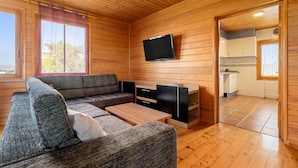 Interior de una cabaña de madera con sofá, televisión, mesa y zona de cocina.