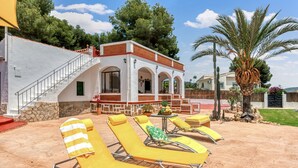 Un edificio con fachada blanca, una escalera y un patio cubierto con sillones amarillos y una mesa.