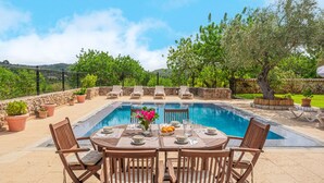 A poolside dining area with a table set for six, surrounded by lounge chairs, and a view of the pool and greenery.