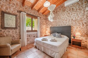 A bedroom with a stone wall, a bed with white linens, a wooden chair, a nightstand with a lamp, and a window with curtains. - Sa Sort Alaró – Charming House with Pool and Mountain Views (Alaró)