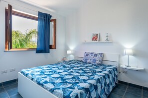 A bedroom with a bed, a window with curtains, a bedside table with a lamp, and a wall-mounted shelf with decorative items.