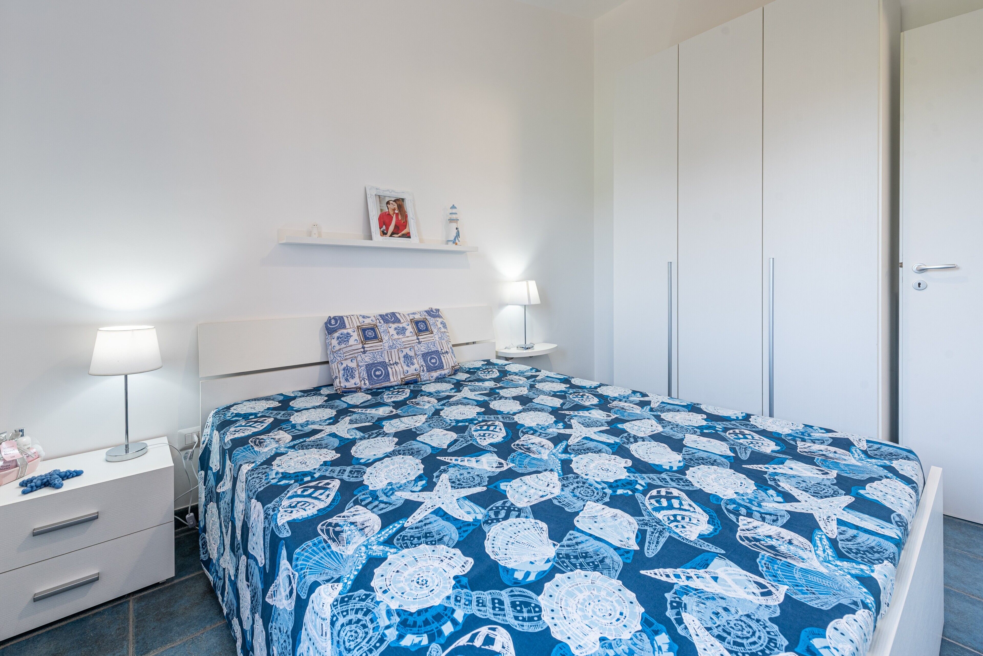 Una camera da letto con un letto, comodini, una lampada, uno scaffale con una foto e una piccola statuina, e un armadio.