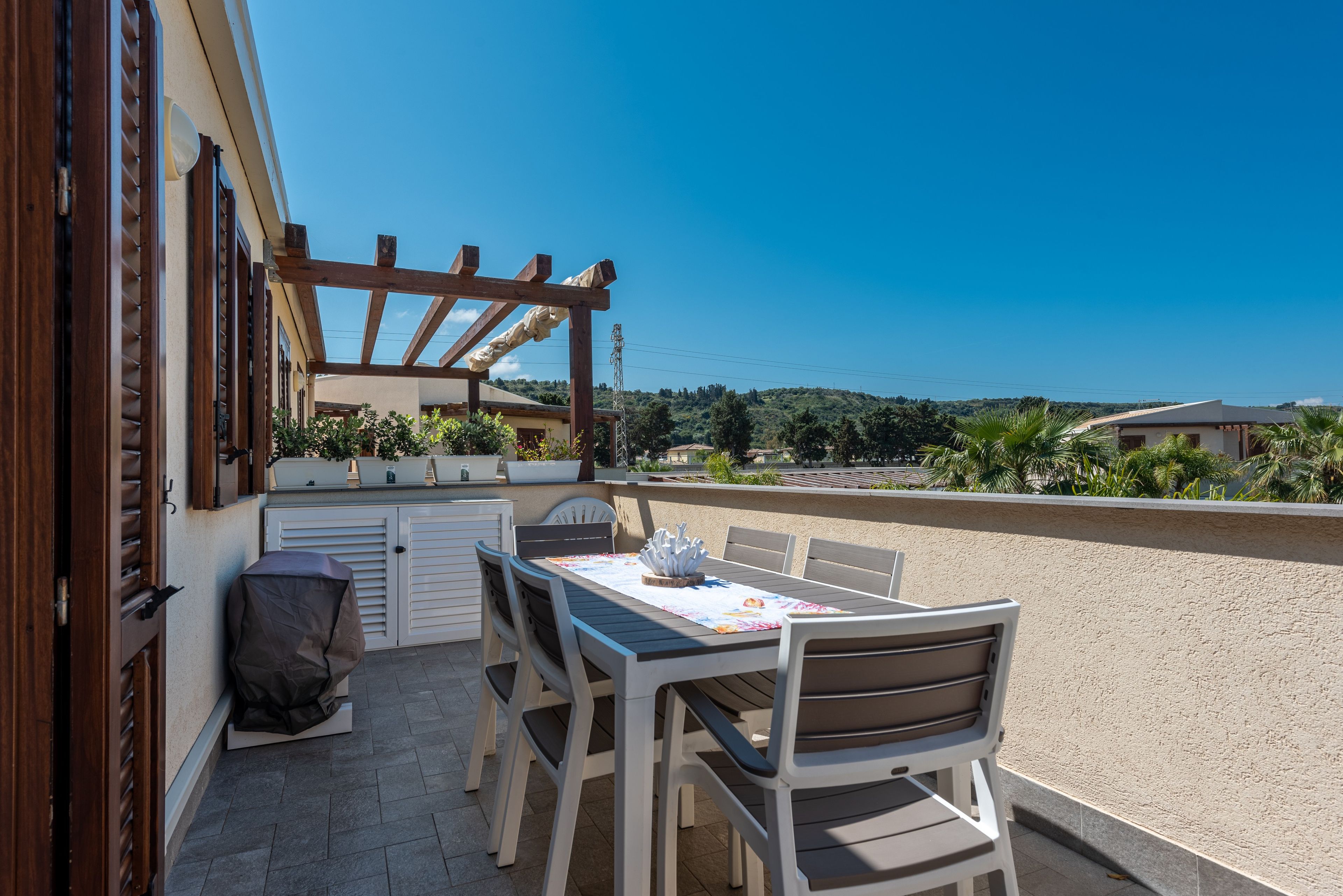 An outdoor patio with a table and chairs, a covered grill, and a view of the surrounding landscape.