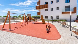 Un parque infantil con columpios y estructuras de escalada, junto a un edificio moderno con balcones.