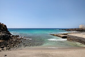Una playa costera con agua cristalina, costa rocosa y un edificio a la derecha.