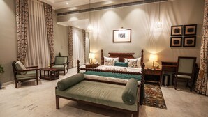 Ein Hotelzimmer mit einem großen Bett, einer Sitzecke mit einer Bank, einem Schreibtisch und einem Stuhl. Vorhänge, ein Bild an der Wand und eine Lampe auf dem Schreibtisch sind ebenfalls vorhanden.