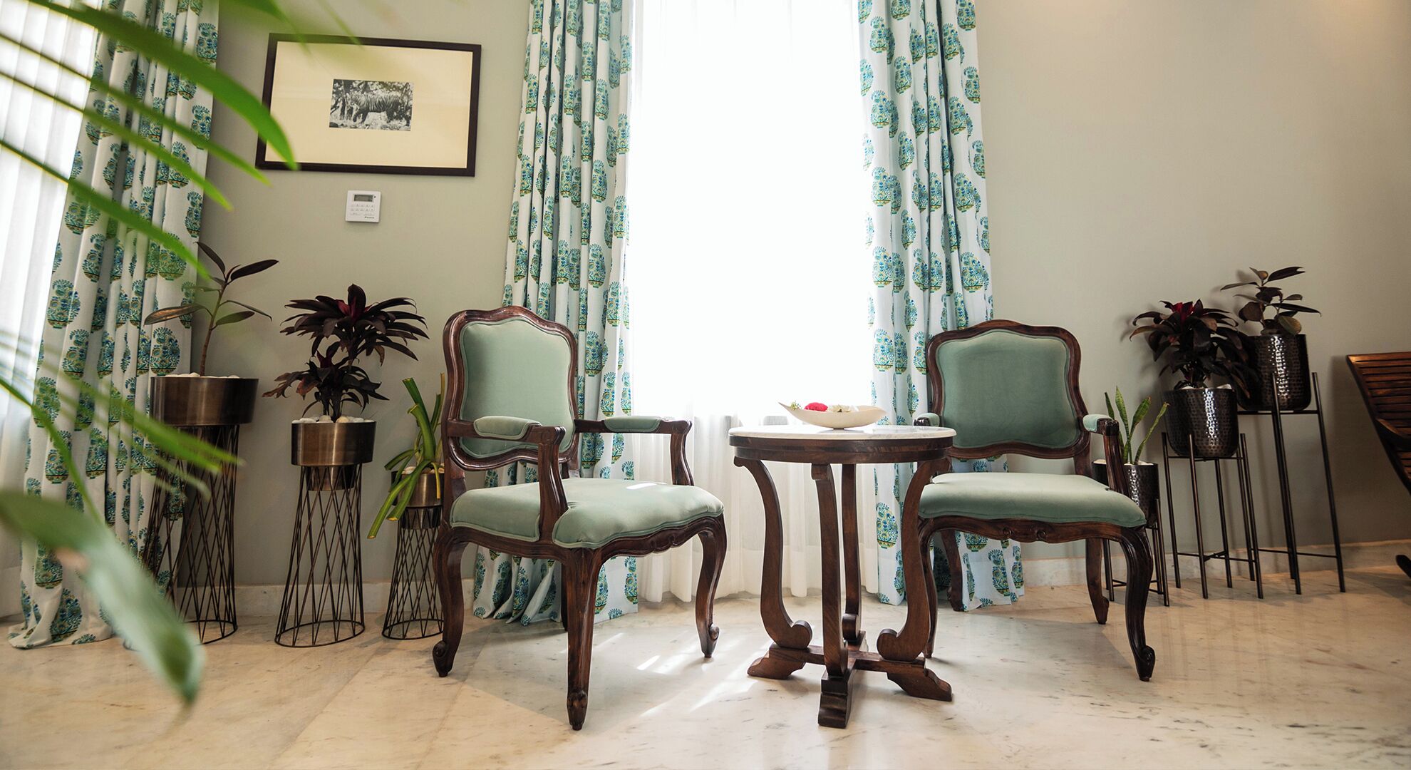 Une pièce comprenant deux chaises en bois et une petite table ronde, le tout agrémenté de rideaux à motifs floraux.