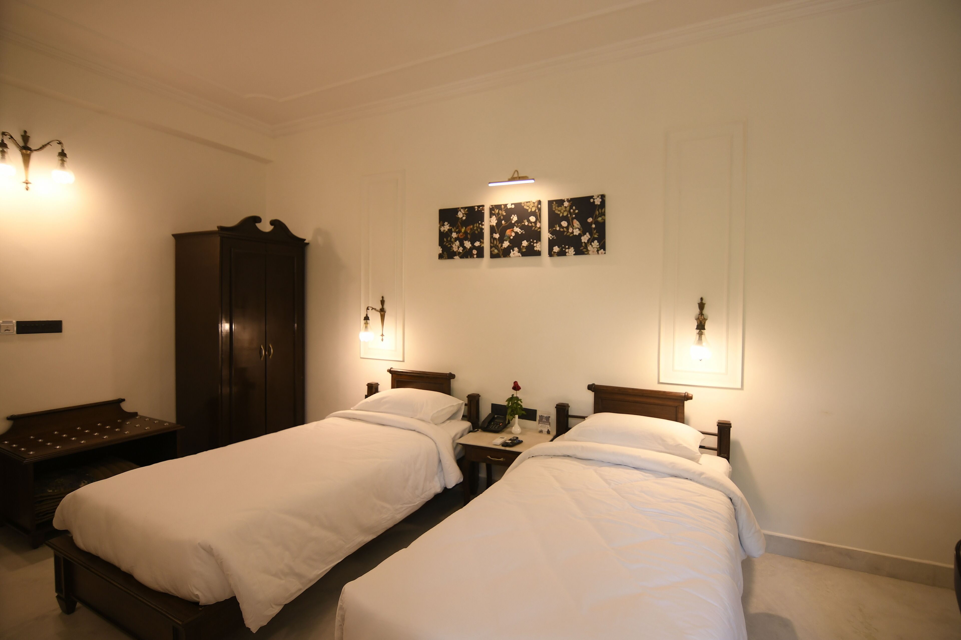 Habitación de hotel con dos camas individuales, un armario de madera, un escritorio con teléfono y un jarrón de flores, además de lámparas fijadas en la pared.