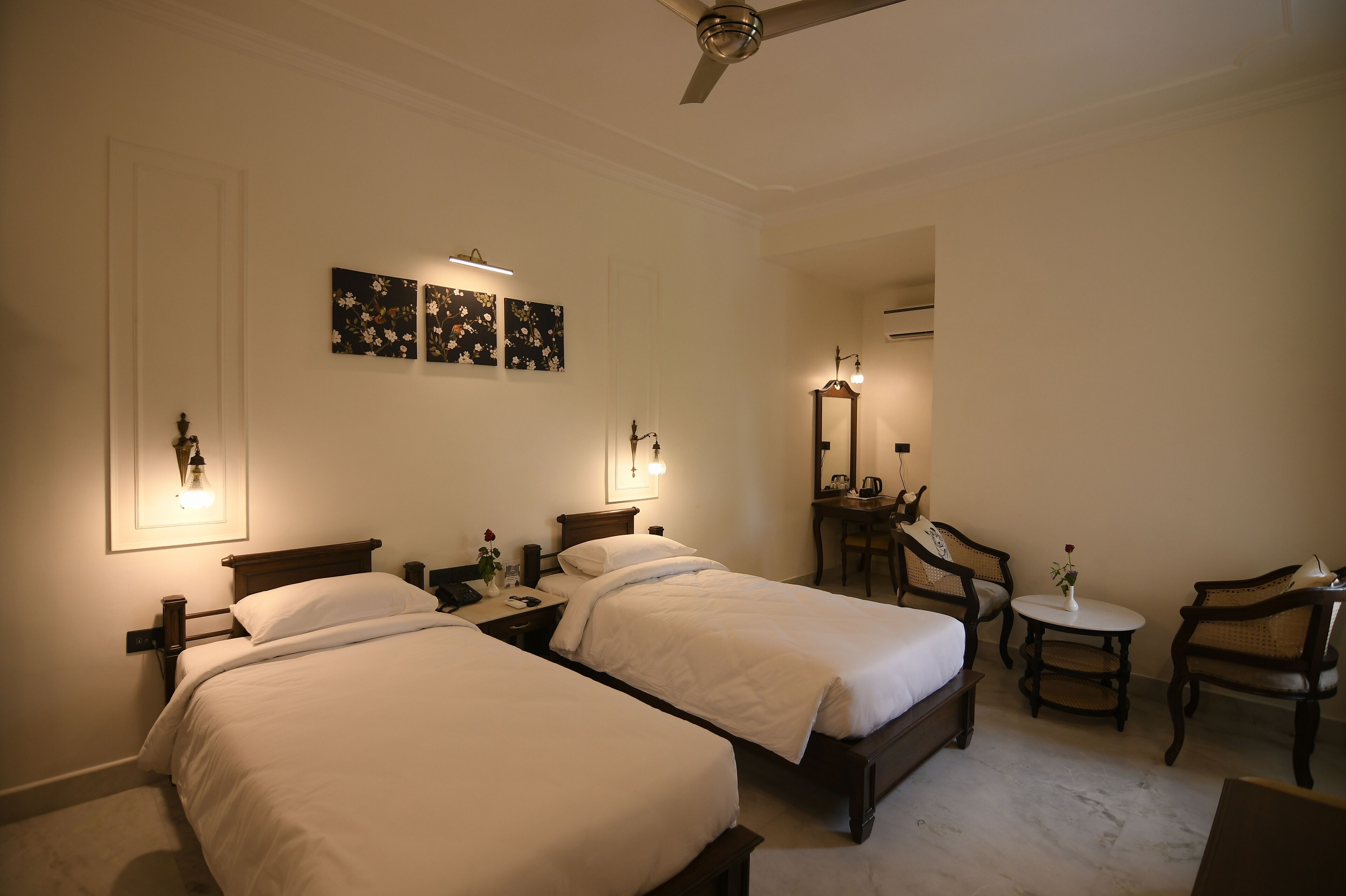 A hotel room with two single beds, a desk, a chair, a small table, and a ceiling fan.