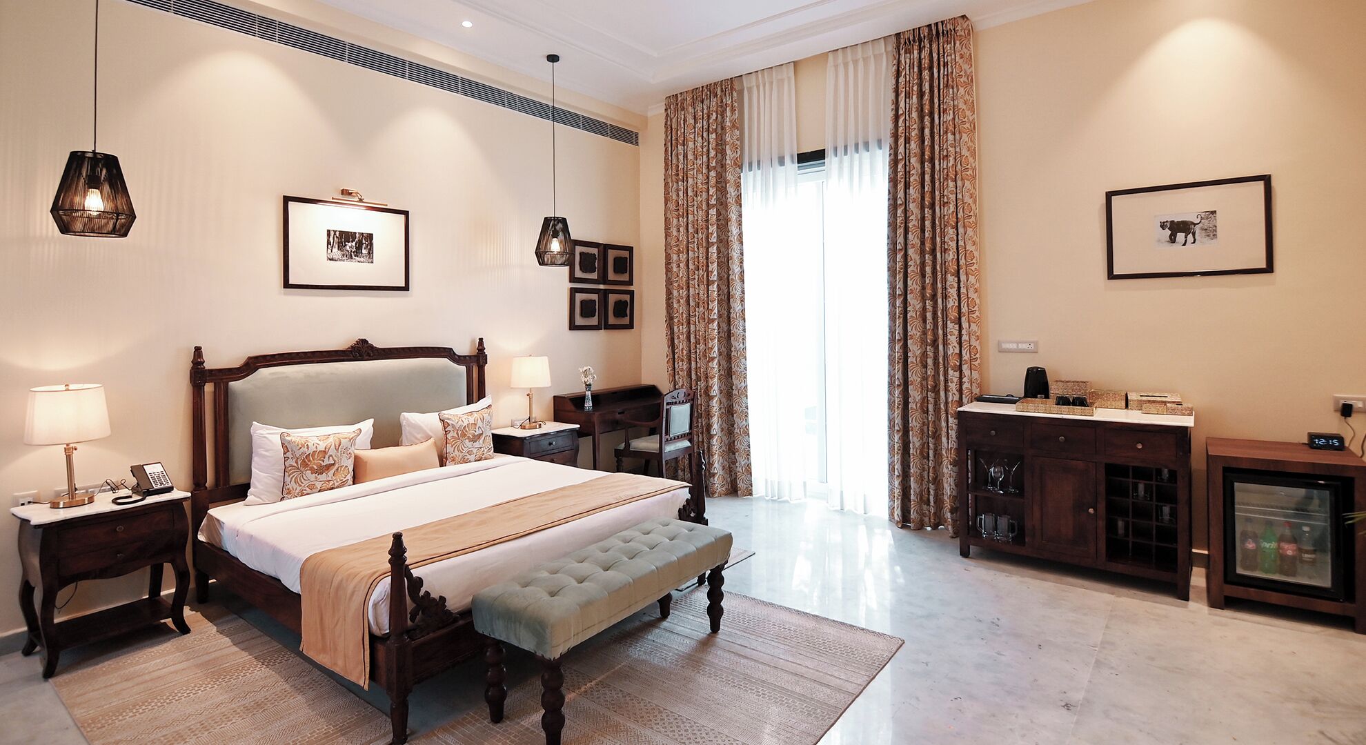 A hotel room with a large bed, bedside tables, a desk, a chair, a minibar, and a large window with curtains.