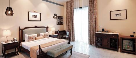 A hotel room with a large bed, bedside tables, a desk, a chair, a minibar, and a large window with curtains.