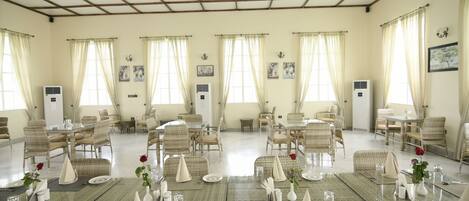 Un vaste espace dédié à la salle à manger, avec plusieurs tables dressées, des chaises en osier et de grandes fenêtres dotées de rideaux.