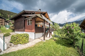 A wooden chalet with a balcony, surrounded by a garden and a mountain backdrop.