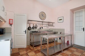 A kitchen with a stainless steel table, hanging pots, a clock, and a fire extinguisher.