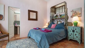 Une chambre à coucher comprenant un lit, une table de chevet, une chaise, un miroir et une salle de bain visible à travers une porte ouverte.