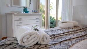 Ein ordentlich bezogenes Bett mit aufgerollten weißen Handtüchern, einer gemusterten Decke und einer weißen Kommode mit Spiegel und einem türkisfarbenen Krug.